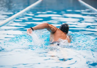 Vīrietis ar glikozes sensoru peldē baseinā, valkājot peldcepuri un sporta pulksteni.