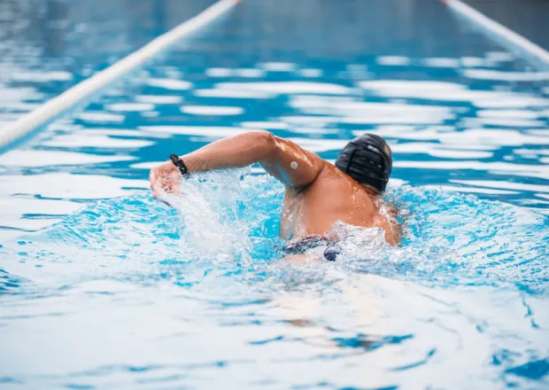 Vīrietis ar glikozes sensoru peldē baseinā, valkājot peldcepuri un sporta pulksteni.